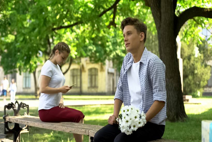 Teenager Hesitating To Present Flowers To Girl In Park Lady Using Smartphone